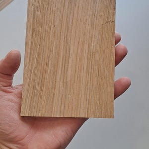 May include: A rectangular piece of light brown oak wood with visible grain patterns. The wood is held in a hand, showcasing its natural color and texture. The wood appears to be a sample or a small piece for a woodworking project.