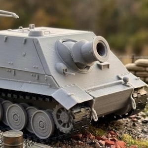 May include: A gray model tank with a large cannon and visible tracks. The tank has a rectangular body with angled sides and a detailed design. The model is set on a terrain of rocks and grass, suggesting a miniature war scene.