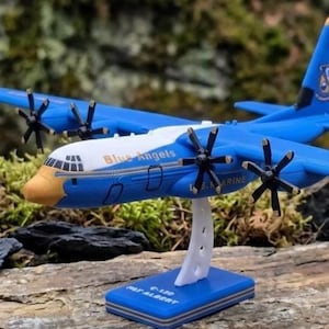 May include: A blue and white model airplane featuring the text Blue Angels and U.S. MARINE on the fuselage. The model has four black propellers, a yellow nose cone, and a vertical stabilizer with a logo. It is mounted on a white stand with a blue base.