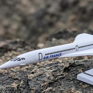 May include: A white model of an Air France Concorde airplane, with the words "AIR FRANCE" printed on the side. The model has a stand with the word "Concorde" on it. The airplane is set against a rocky background.