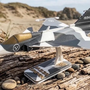 May include: A JAS39 Gripen fighter jet model featuring a gray, white, and black camouflage pattern, a gold-tinted cockpit canopy, and the number 1994 on the tail. The model is mounted on a white stand with the text JAS39 GRIPEN printed on the base.