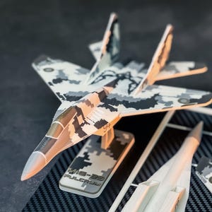 May include: A model of a MiG-29 Fulcrum fighter jet with a camouflage pattern in shades of gray, white, and brown. The model is on a stand with the text "MiG-29 Fulcrum". Additional unassembled parts are visible.