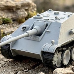 May include: A grey model tank with a long barrel, detailed tracks, and a textured surface. The miniature tank is set against a backdrop of rocks and foliage, suggesting a battlefield setting. The tank's design includes visible hatches and other features.