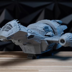 May include: A light gray, unpainted 3D printed model of a spaceship featuring a prominent cockpit, detailed hull paneling, and two side-mounted engine thrusters. The spacecraft has a complex, angular design with a rounded rear section.