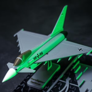 May include: A model airplane with a gray body and green accents, including the wings and fuselage. The model has the number "19+94" and a cross symbol on the side. The plane is resting on a dark surface.