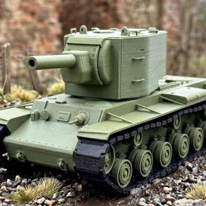 May include: A detailed model of a military tank in olive green. The tank features a long barrel, a rotating turret, and multiple wheels with black treads. The model is set against a blurred outdoor background.