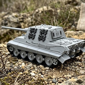 May include: A gray model tank with a long barrel and detailed tracks. The miniature tank is set on a bed of small rocks and dirt, showcasing its design. The tank's features include multiple wheels and intricate detailing.