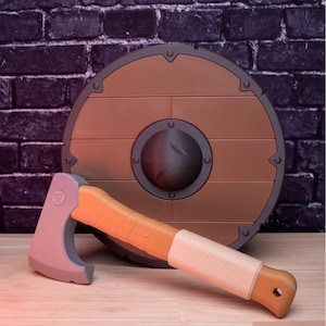 May include: A toy Viking shield and axe set. The shield is round, brown, and has a dark gray center. The axe has a gray head, an orange handle, and a white wrapped grip. The set is on a wooden surface.