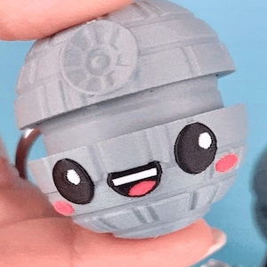 Puede incluir: Un llavero de la Estrella de la Muerte gris con una cara de dibujos animados. El llavero tiene una boca sonriente, ojos negros con reflejos blancos y mejillas rosadas. La Estrella de la Muerte es una esfera con paneles detallados.