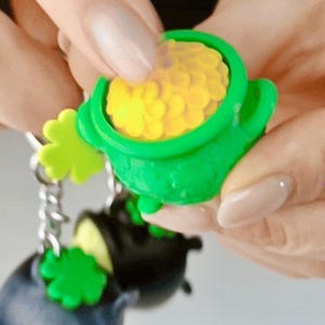 Può includere: Un portachiavi a forma di calderone in plastica verde brillante riempito con monete gialle testurizzate. Il portachiavi presenta un piccolo ciondolo a forma di quadrifoglio verde attaccato a una catenella in metallo argentato e forma tonda.