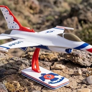Puede incluir: Un modelo de avión blanco y azul con detalles en rojo, incluyendo la cola y la nariz. El modelo tiene "USAF" impreso en el lateral y está montado en un soporte con las palabras "Thunderbirds" y un logotipo.