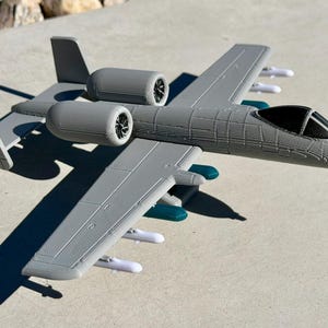 May include: A gray model airplane with two engines, wings, and a cockpit. The model has white missiles attached to the wings and teal accents. The airplane is sitting on a concrete surface.