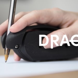 May include: A black 3D-printed ergonomic pen grip holder with a matte finish, featuring a secure clip mechanism holding a standard ballpoint pen. The word DRAG is printed in white capital letters across the side of the device.