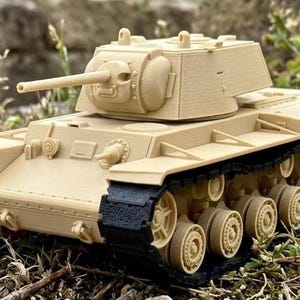 Op de afbeelding: Een tan-kleurig model tank met zwarte rupsbanden en wielen. De tank heeft een lange loop en een gedetailleerde toren. Het model staat tegen een achtergrond van groen, dat lijkt op een replica van een militair voertuig.