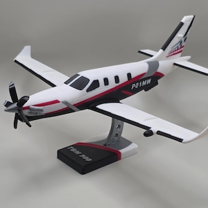May include: A scale model of a TBM 900 airplane featuring a white, black, and red color scheme. The fuselage is marked with the text P01MW, and the black display base is labeled TBM 900. The aircraft has a five-blade propeller and winglets.