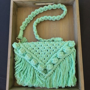 May include: A mint green macrame crossbody bag with a woven design and fringe detail. The bag features a braided strap and decorative knots. The bag is displayed in a wooden tray.
