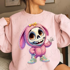 Puede incluir: Sudadera rosa claro con un personaje de esqueleto de dibujos animados con un disfraz de conejito rosa y un lazo amarillo. El personaje tiene ojos marrones grandes y lleva zapatillas deportivas coloridas. El diseño es caprichoso y juguetón.