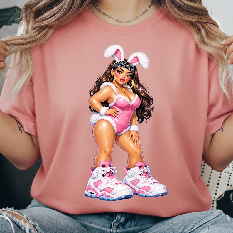 Puede incluir: Una camiseta rosa polvoriento con una ilustraci&oacute;n gr&aacute;fica de un personaje de pelo largo casta&ntilde;o que viste un body rosa, orejas de conejo blancas y zapatillas altas blancas y rosas. El personaje lleva un pa&ntilde;uelo negro y pendientes de aro dorados.