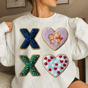 May include: White sweatshirt with embroidered designs. Features a blue X, a green X, and two heart-shaped patches. One heart has two teddy bears, and the other has red and white hearts on a blue background. Gold trim.