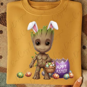 Pode incluir: Camisola mostarda com um personagem de desenho animado Groot com orelhas de coelho, segurando uma cesta de ovos de Páscoa. Uma casca de ovo roxa com o texto "Happy Easter" também está presente, juntamente com ovos de Páscoa coloridos.