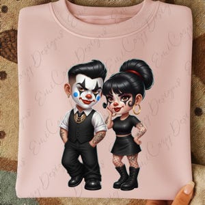May include: A light pink sweatshirt featuring a cartoon couple dressed as clowns. The male clown wears a black suit, white shirt, and face paint. The female clown wears a black outfit and hoop earrings.