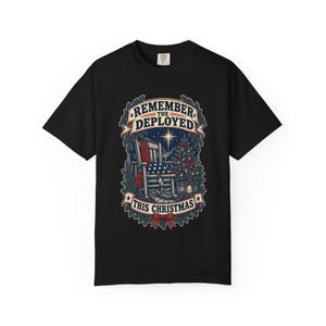 May include: A black short-sleeved t-shirt featuring a graphic print of a wooden rocking chair with an American flag draped over it, a decorated Christmas tree, and a lantern. The text reads REMEMBER THE DEPLOYED THIS CHRISTMAS in a banner.