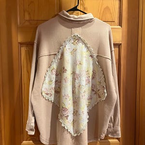 May include: A tan long-sleeved shirt with a floral patterned panel on the back. The panel is a diamond shape with a white lace trim. The shirt is hanging on a black hanger.