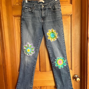 Op de afbeelding: Blauwe spijkerbroek met drie grote, kleurrijke bloemenapplicaties op de voorkant. De bloemen hebben tinten geel, groen, roze en paars. De jeans hangt aan een doorzichtige hanger.