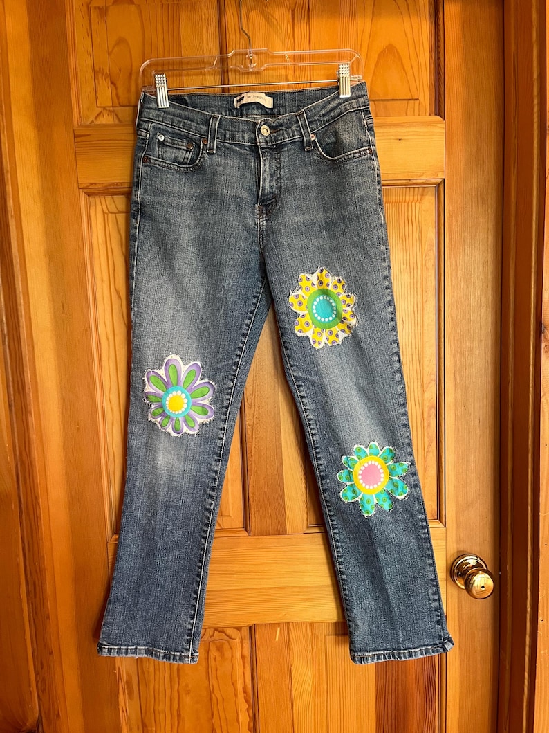 Op de afbeelding: Blauwe spijkerbroek met drie grote, kleurrijke bloemenapplicaties op de voorkant. De bloemen hebben tinten geel, groen, roze en paars. De jeans hangt aan een doorzichtige hanger.