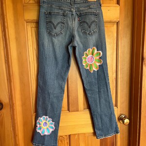 Op de afbeelding: Blauwe spijkerbroek met twee grote, kleurrijke bloemdesigns op de benen. De bloemen hebben roze, oranje, groene en blauwe bloemblaadjes. De jeans heeft een klassiek ontwerp met vijf zakken.