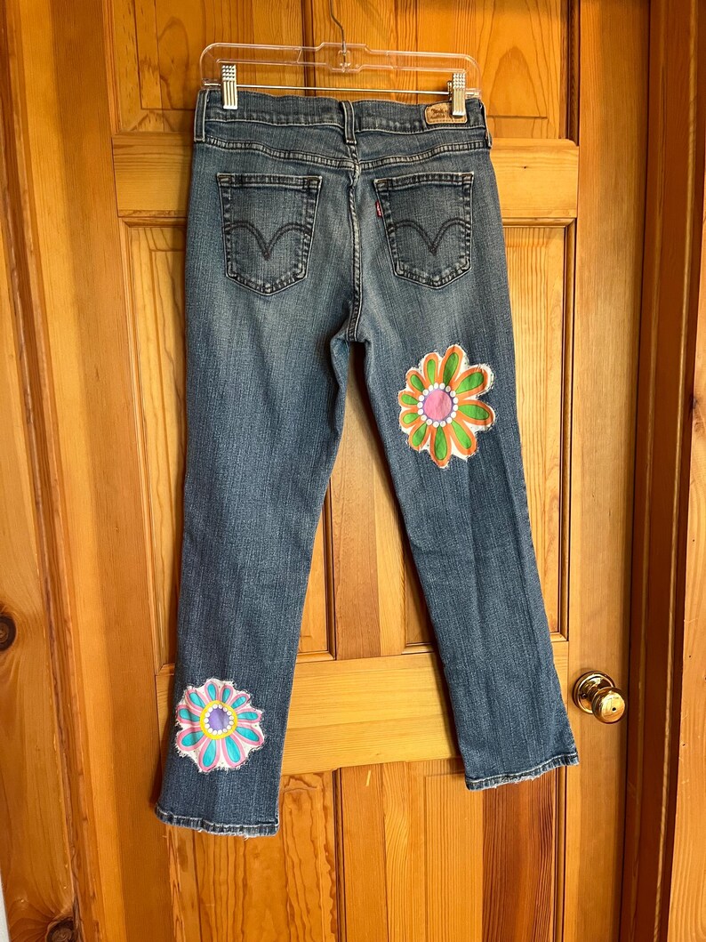 Op de afbeelding: Blauwe spijkerbroek met twee grote, kleurrijke bloemdesigns op de benen. De bloemen hebben roze, oranje, groene en blauwe bloemblaadjes. De jeans heeft een klassiek ontwerp met vijf zakken.