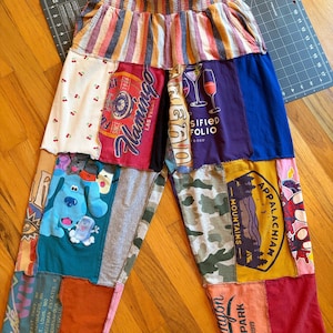 May include: Patchwork pants featuring a striped elastic waistband and various graphic fabric panels. Prints include a blue dog character, a Las Vegas Flamingo logo, a wine glass illustration, an Appalachian Mountains emblem, and a Canyon National Park graphic.