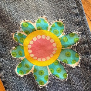 Op de afbeelding: Close-up van een blauwe spijkerbroek met een grote, kleurrijke bloem patch. De bloem heeft een gele kern met een roze binnenste cirkel en witte stippen. De bloemblaadjes zijn turquoise met groene stippen.