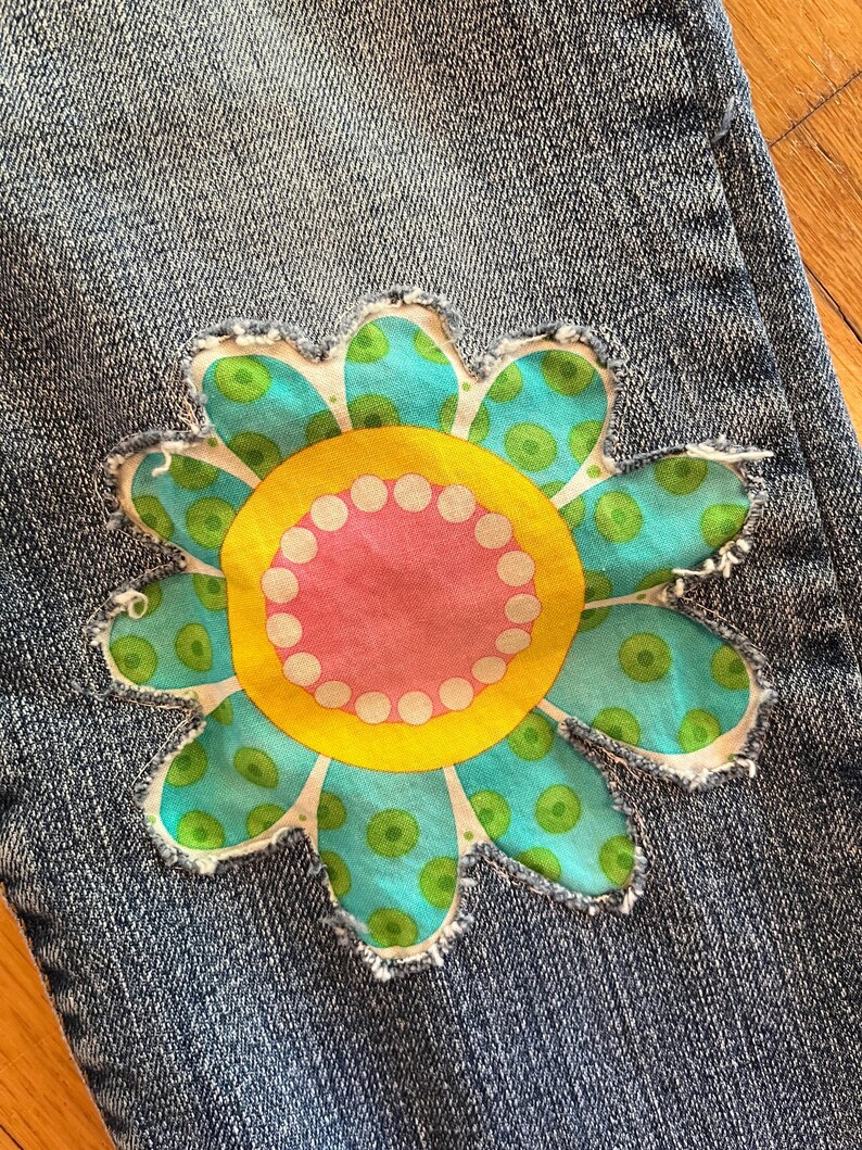 Op de afbeelding: Close-up van een blauwe spijkerbroek met een grote, kleurrijke bloem patch. De bloem heeft een gele kern met een roze binnenste cirkel en witte stippen. De bloemblaadjes zijn turquoise met groene stippen.
