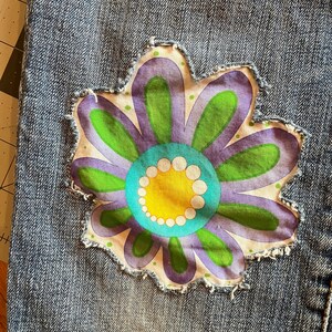 Op de afbeelding: Close-up van een denim stof met een grote, kleurrijke bloem patch. De bloem heeft paarse, groene, gele en turquoise tinten, met een wit stippenpatroon. De patch heeft een gerafelde rand, wat een vintage touch geeft.