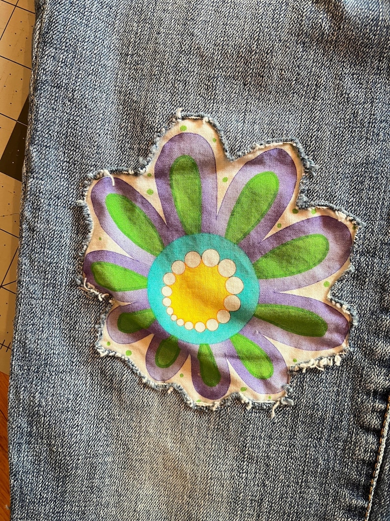 Op de afbeelding: Close-up van een denim stof met een grote, kleurrijke bloem patch. De bloem heeft paarse, groene, gele en turquoise tinten, met een wit stippenpatroon. De patch heeft een gerafelde rand, wat een vintage touch geeft.