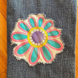 Op de afbeelding: Close-up van blauwe denimstof met een grote, kleurrijke bloemenpatch. De bloem heeft roze, teal, gele en paarse details. De patch heeft een gerafelde rand, wat een vintage of handgemaakte uitstraling geeft.