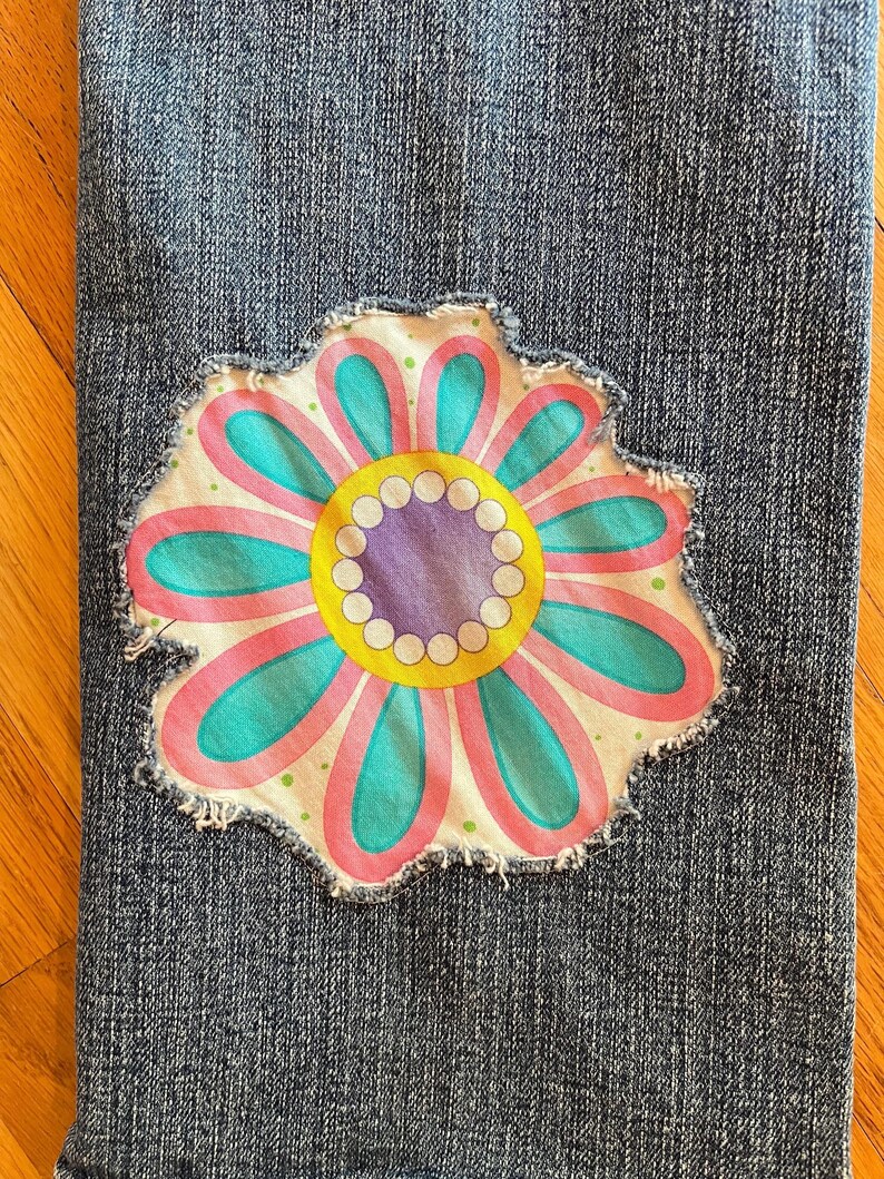 Op de afbeelding: Close-up van blauwe denimstof met een grote, kleurrijke bloemenpatch. De bloem heeft roze, teal, gele en paarse details. De patch heeft een gerafelde rand, wat een vintage of handgemaakte uitstraling geeft.