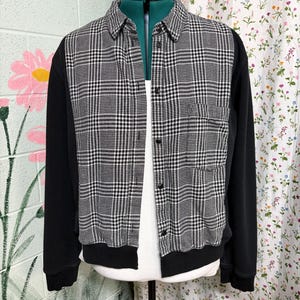 May include: A long-sleeve button-up shirt featuring a black and white glen plaid pattern on the front panels and a solid black fabric on the sleeves and back. The shirt includes a pointed collar, a single chest pocket, and black buttons.