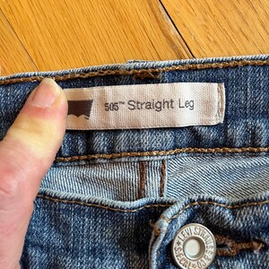 Op de afbeelding: Close-up van een blauwe spijkerbroek. Het label van Levi Strauss & Co. luidt "505 Straight Leg". Een hand houdt het label vast. De jeans heeft een knoop met de woorden "LEVI STRAUSS & CO." en "SEGAL 03".