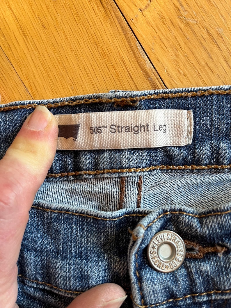 Op de afbeelding: Close-up van een blauwe spijkerbroek. Het label van Levi Strauss & Co. luidt "505 Straight Leg". Een hand houdt het label vast. De jeans heeft een knoop met de woorden "LEVI STRAUSS & CO." en "SEGAL 03".