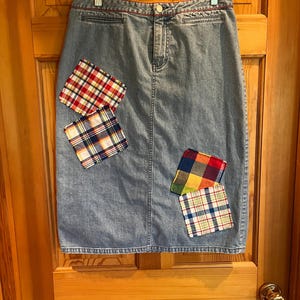 May include: A light-wash denim skirt with a straight cut, featuring a red stitched waistband and decorative plaid patches in red, blue, and yellow. The skirt hangs on a clear hanger against a wooden door.