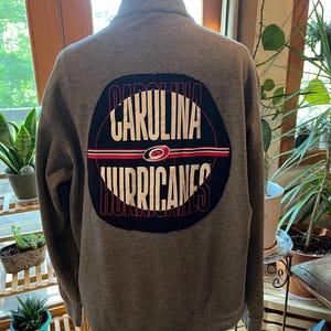 Puede incluir: Una sudadera gris con cremallera y un parche circular grande en la espalda. El parche presenta las palabras "CAROLINA HURRICANES" en blanco, rojo y negro, con el logotipo del equipo en el centro.