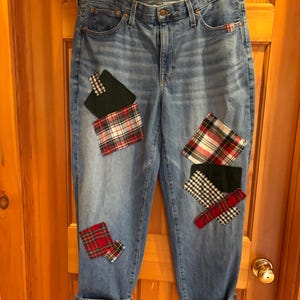 May include: Medium-wash blue denim jeans featuring a high-rise waist, a button fly, and multiple fabric patches in red, white, and black plaid, green, and checkered patterns sewn onto the front legs. The jeans have cuffed hems.