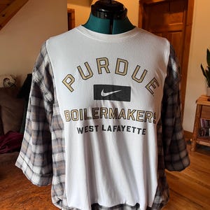 May include: A white crewneck sweatshirt with plaid sleeves and a lower hem. The front features the text PURDUE in gold, a black Nike logo, and the text BOILERMAKERS WEST LAFAYETTE in black and gold lettering on a white background.
