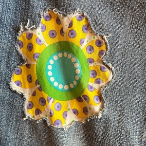 Op de afbeelding: Een close-up van denimstof met een bloemenpatch. De bloem heeft gele bloemblaadjes met paarse stippen, een groene kern en een turquoise kern met witte stippen. De patch heeft een gerafelde rand.