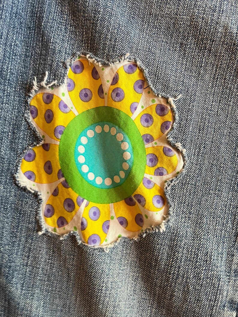 Op de afbeelding: Een close-up van denimstof met een bloemenpatch. De bloem heeft gele bloemblaadjes met paarse stippen, een groene kern en een turquoise kern met witte stippen. De patch heeft een gerafelde rand.