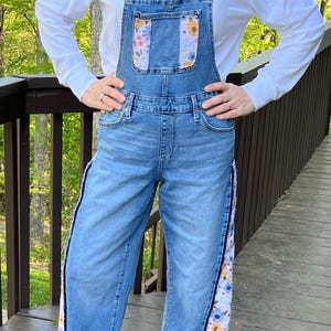 May include: Light blue denim overalls with floral fabric accents on the bib pocket and side panels. The overalls have adjustable straps and a relaxed fit. The floral fabric features yellow, blue, and pink flowers. The bottom hem is frayed.