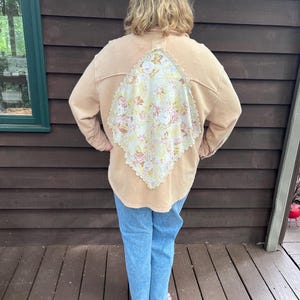 May include: A tan long-sleeved shirt featuring a large diamond-shaped panel on the back made of white fabric with a yellow and pink floral pattern, bordered by cream-colored lace trim, paired with light blue denim jeans.