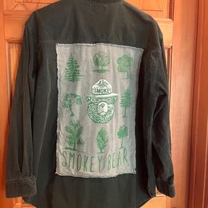 May include: Dark green button-down shirt with a Smokey Bear graphic on the back. The graphic features Smokey Bear's face, trees, and the words "Smokey Bear" in green on a light green patch.